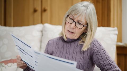 A woman with grey-blonde hair wears a lilac jumper and glasses as she sits on a sofa looking at bills 