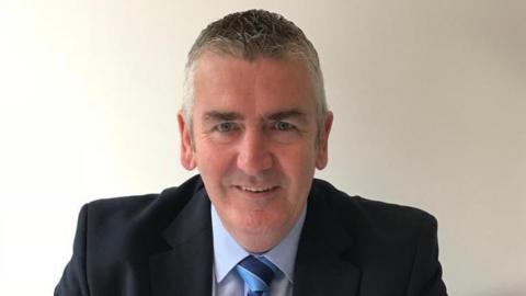 David McWilliams pictured sat against a white wall. He is wearing a blue shirt and blue tie.