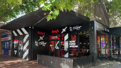 A shop front which has black signage with red and white writing that reads Betsy's X Little Dessert Shop. There are trees in front of the shop.