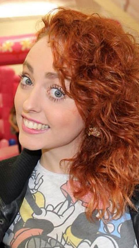 A young girl with red hair smiling at the camera with a colourful t-shirt on.