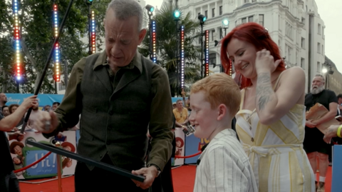Tom Hanks is standing on a red carpet and holding and looking at the framed winning design. Tommy is also looking down at the painting and smiling. Crowds of people can be seen around them, and the edge of some Toy Story 5 branding can be seen behind Tommy and a woman with red hair who is standing next to him and smiling.
