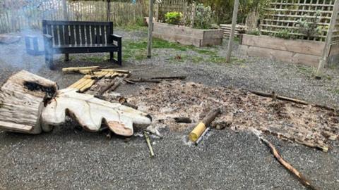 Part of the garden showing a smoking leaf sculpture on its side to the left and a burning patch of scorched earth and wood to the right