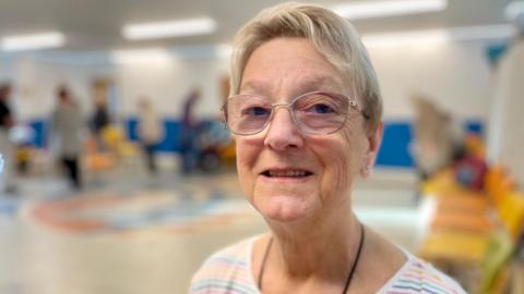 Stephanie smiles for the camera wearing a white jumper with people in a room behind her 