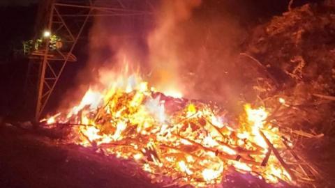 A picture of a bonfire. It looks to be burning on the side of a major road, with a pylon visible in the background.