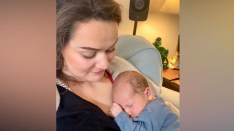 A mother is holding her son who is a newborn. The baby is lying into her chest and is asleep. She is looking at him tenderly. 