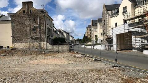 The former CI Tyres site is level and gravelled. There is a metal fence around it. There are houses next to and opposite it.