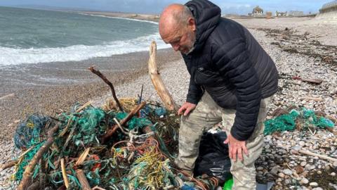 A man leans over a pile of tangled fishing rope and nets on a beach