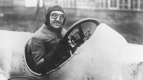 A black and white photo of an early 20th Century racing driver. He is sitting inside a car, wearing a pair of goggles and protective headwear.