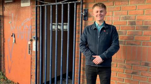 A young man stands in front of a public toilet – this is a red brick building with a metal fence-style door. He is smiling at the camera.
