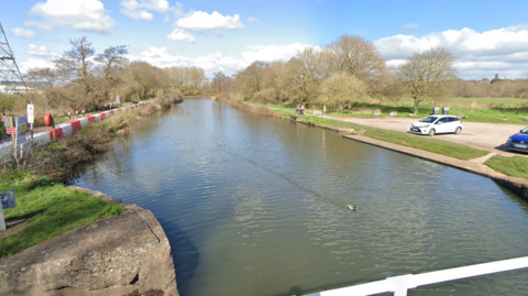 Google street view image of Clapperbrook Lane East overlooking the canal. It is a sunny day, the photo has been taken from the bridge that overlooks the water.