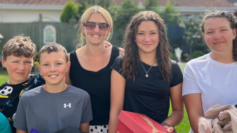 (left to right James (10) , Henry (11), Mum Nikki, Grace (14) and Olivia (16)). They are all smiling holding their back to school purchases and hand-me downs. The family is standing in the garden on a sunny day. They all have black hair apart from the mum who is blonde.