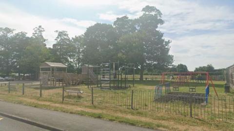 An outdoor play park with multiple pieces of play furniture, including swings and climbing frames. Trees sit behind it. The sky is blue and cloudy.