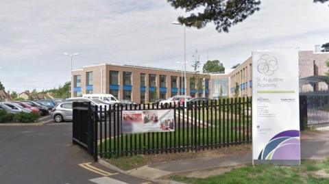 A Google maps image showing a sign for St Augustine Academy and the large v-shaped school building in the background beyond a green fence and a carpark