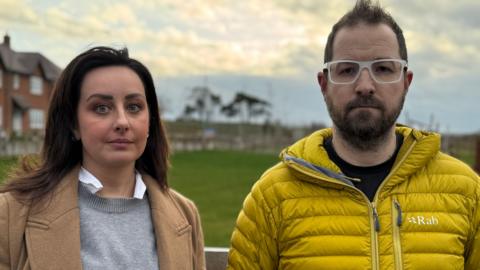A man wearing white glasses and a yellow coat and a woman wearing a grey jumper with a brown coat are pictured from the shoulders up, stood in front of a grassy area with houses in the background.
