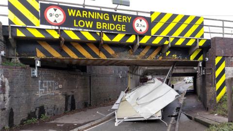 A low bridge with 'Warning very low bridge' written on top with bright yellow and black stripes behind it. There is also a sign showing the height as 2.7m. Under the bridge a white van has become stuck and the roof has been ripped off it. 