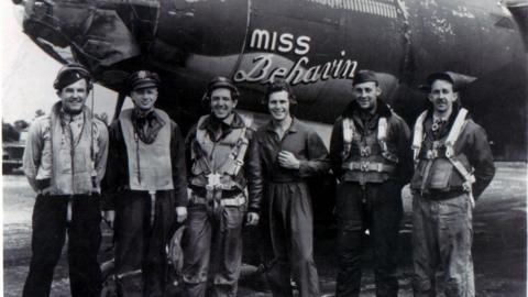 A black and white image of six men standing in front of an aircraft named "Miss Behavin". They are smiling.