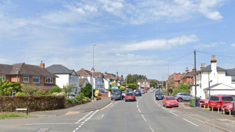 A main road with a side road on the left. Cars are seen parked and driving on both sides of the road, with a row of houses and a street sign in the background.