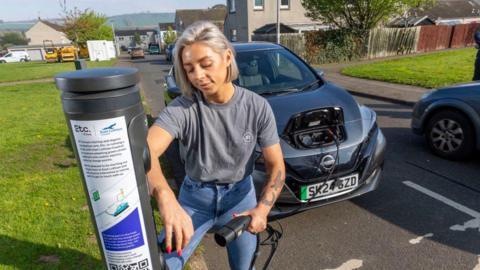 A woman plugs her electric car into the public charging point on the street in a housing estate.