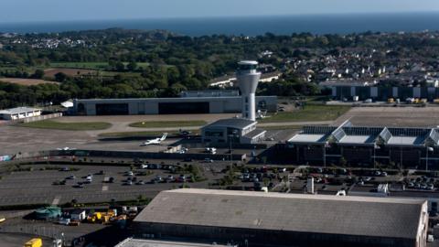 An aerial photo of Jersey Airport on a bright day which shows the car parks, air traffic control tower and hangars. In the distance there are trees and the sea.