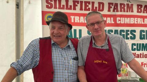 Peter Gott and a colleague standing in a market stall as they sell sausages. They are dressed in chequered shirts and red aprons. Peter is wearing a brown hat.
