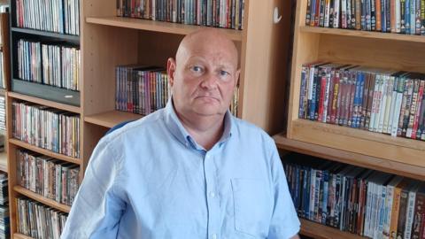 Mark is wearing a light blue shirt and is sitting in front of a wall of rare Dvds on light brown shelves.