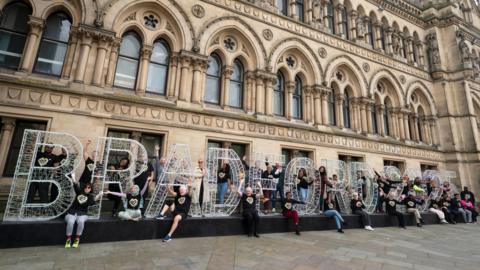 A group of people in black T shirts stand around 6-8ft letters that read 'Bradford' in front of Bradford City Hall. 