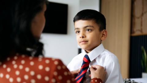A woman is putting on her young boy's red and blue stripped tie for school. She is wearing an orange and white polka dot blouse and has brown hair her back is to the camera is blurred. The boy has brown hair and is wearing a white shirt