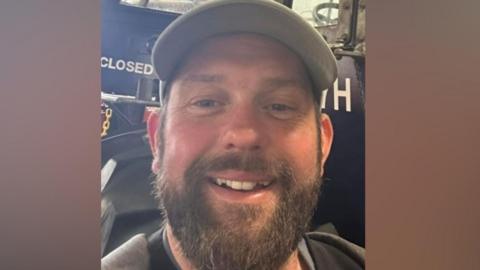James Desborough is smiling. He has a brown beard and is standing in front of a blue tractor. He has a grey baseball cap on.