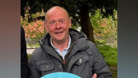 Tierney is stood outside by some trees and is smiling at the camera. He has a grey jacket on over a grey shirt. He has short brown hair.