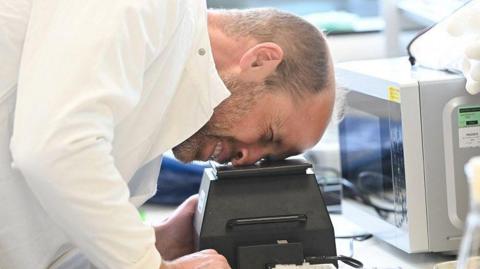 Prince William looking at something through a machine while dressed in a lab coat. 