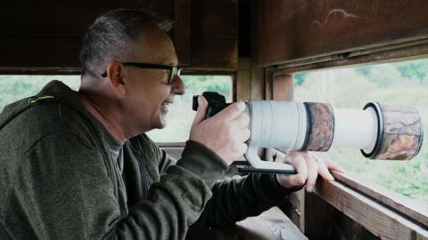 Alan Bruce is sitting in a wooden wildlife hide. He has a long-lens camera resting in front of him, sticking out of the open side of the hide. Alan is wearing a green hoodie and has dark-rimmed glasses