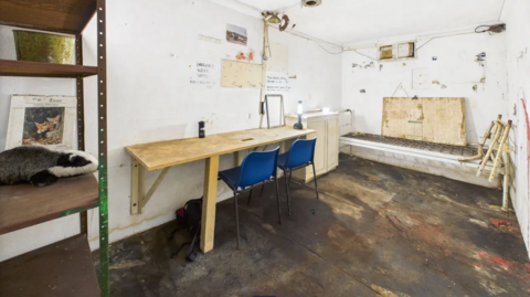 Interior of the bunker which has white walls a small wooden desk and two blue plastic chairs. At the near left side, there is a basic-looking stack of shelves which are  brown and stained.