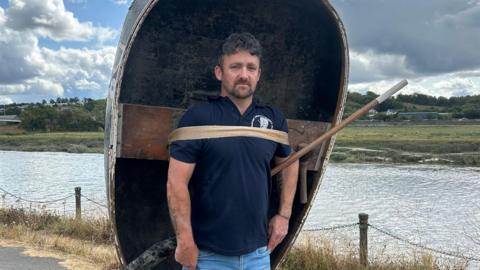 Leighton Morgans mewn crys polo glas tywyll a jeans glas golau. Yn sefyll gyda chwrwgl; cwch bach crwn ar ei gefn yn sefyll ar lan yr afon Tywi.