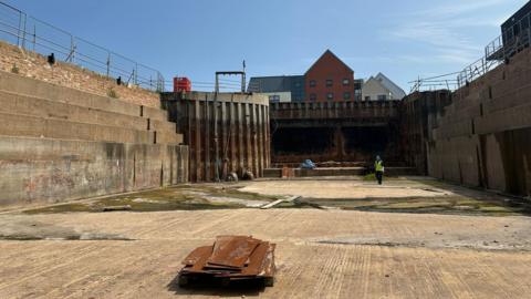 North Bridge Dry Dock from inside the dock. A person in black trousers, a black t-shirt, a yellow hi-visibility vest and a blue hard hat can be seen in the distance. The sky is blue.