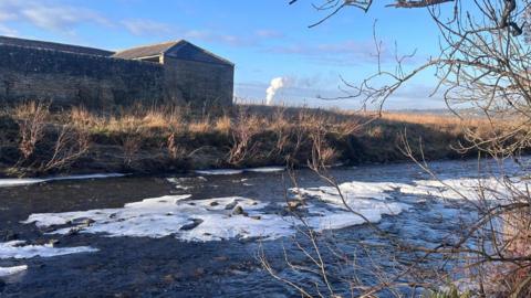 Floating sheets of ice have formed along the top of a river. They have formed around the shallow rocks parts in the middle of the river. A stone building can be seen on the far bank.