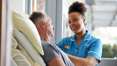 A Sue Ryder nurse smiling down at a male patient who is lying on a bed