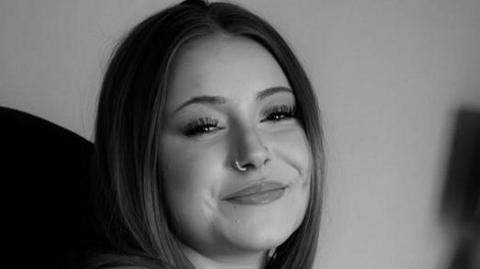A woman smiles at the camera in a black and white image. She has a nose ring in her right nostril. Her hair is parted in the middle