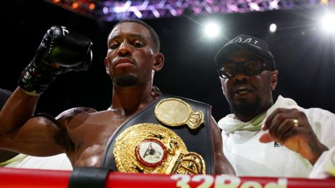 Ezra Taylor with a boxing belt over his shoulder is stood beside coach Malik Scott in the ring