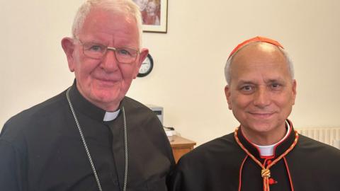 Bishop Campbell (left) has grey hair, metallic spectacles and a silver chain and is wearing a white clerical dog collar with a black shirt. Bishop Robert Prevost, now Pope Leo XIV is wearing a black cardinal's soutane with red edgings and buttons, and a red skull cap. He has thin greying hair.
