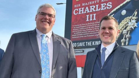 Gavin Robinson stands beside Robert Jenrick as the two smile in a photo together. Robinson is a middle aged man with short, white hair and he is wearing glasses as well as a light gray suit jacket, a white shirt and a blue patterned tie. On his right is Jenrick, who is wearing a zip up hoodie underneath a dark grey suit jacket. The two are stood in front of a mural of King Charles III.