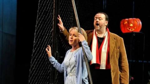 A man in brown coat and red and white scarf, stands and sings behind a woman who is in a blue cardigan and grey dress.