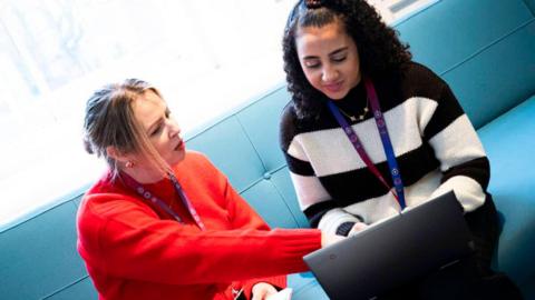 A woman in an orange jumper and a girl in a black and white striped jumper sit on a blue sofa looking at a laptop. The woman in orange is pointing at something on the laptop screen. 