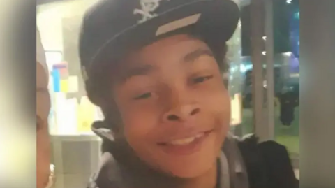 a young boy wearing a dark cap and jacket smiles at the camera