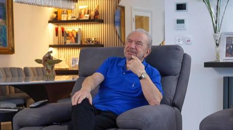 Lord Sugar wears a blue polo shirt and sits in a grey armchair