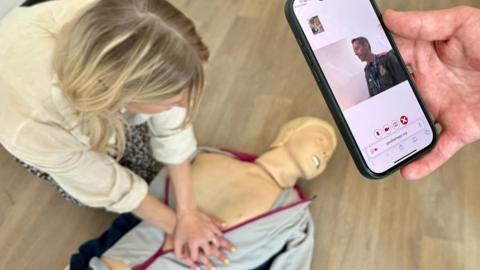 A woman attempts CPR on a sim dummy used to simulate emergency situations. The dummy is dressed in a zip up tracksuit top and has its head turned to one side. You can see its teeth. The woman has her arms out straight in front of her and has clasped one hand on top of the other on the dummy's chest. She has blonde hair and is looking at his chest to check for breaths. A phone is held to the right of the screen by a second person looking at a video call. It shows an advanced paramedic from the East of England Ambulance Service sat at a computer, guiding her on what to do. 