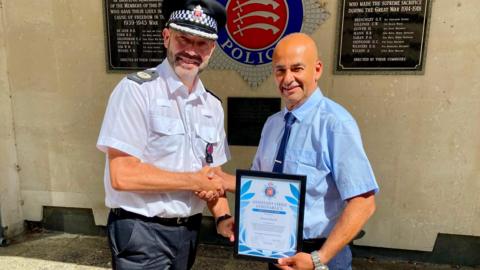 Glen Pavelin is wearing his police uniform and is shaking El Kamel's hand. El Kamel is dressed in a blue shirt and navy tie and is holding the certificate as the award is presented.