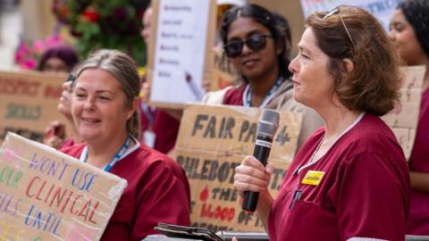 A group of people on a picket line. They are wearing a dark red medical uniform and holding placards reading 'Fair Pay' and 'Respect Our Skills'. One women toward the the front of the image is holding a microphone and looks like she is about to talk. She has dark hair and sunglasses on her head. 