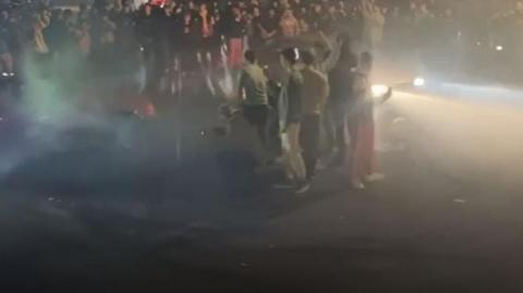 Two cars with headlights on driving around a circle of people at night. You can see the cars' lights beaming into the crowd of people.