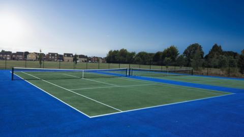 Two hard tennis courts with revamped surfaces. The courts are dark green and have a royal blue perimeter. Outside of the fenced courts is a public park with grass, surrounded by trees and houses. It is a sunny day.