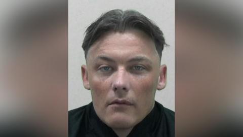 Mugshot of Morgan. He has thick dark and grey hair shaved at sides and long on top in a centre parting. He is clean shaven with a glazed expression.
